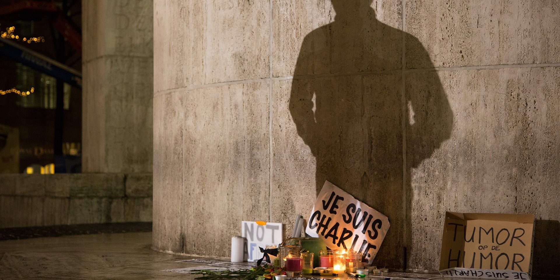 protest-1273818_1920 Kwiaty i znicz oraz karton z napisem: je suis charlie, postawione przy fasadzie budynku.